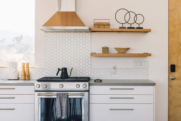 A modern kitchen with a gas stove, oven, and a wooden range hood. There are white cabinets, a hexagonal tile backsplash, and wooden shelves with decorative items. - Hawk & Mesa By Homestead Modern (Pioneertown)