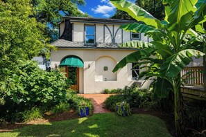 Une maison à deux étages avec un porche à l’avant, un petit jardin agrémenté de plantes en pot et un grand bananier au premier plan.