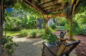 A covered outdoor seating area with a pergola, a table, and chairs, surrounded by lush greenery and a house in the background. - Serene Cottage with Lush Landscaped Grounds (Charleston)