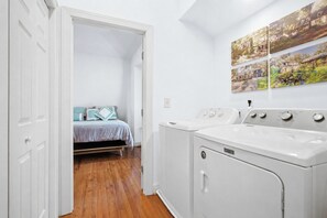 A laundry room with a front-loading Maytag washer and dryer, a bed with a blue and white bedspread, and a hallway leading to a bedroom.