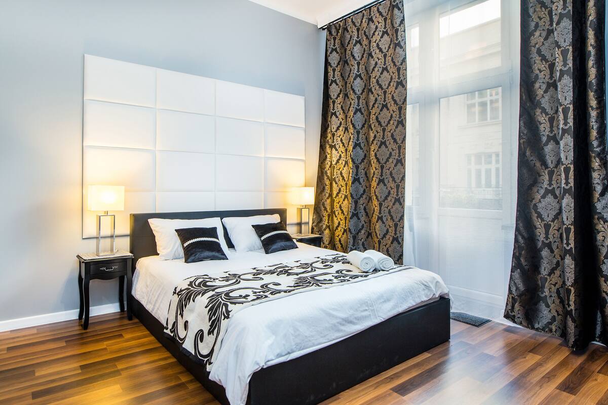 A modern bedroom with a large bed, two bedside tables, and a window with patterned curtains.