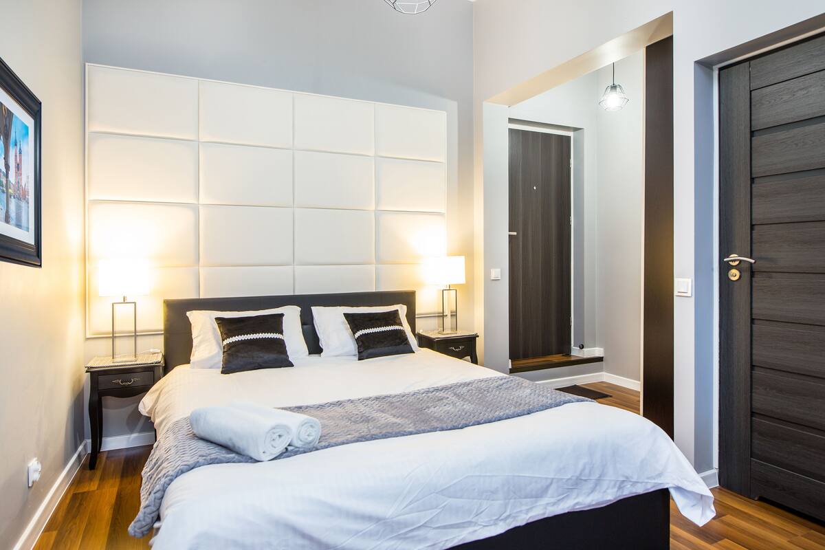 A neatly made bed with a white comforter and two pillows, a bedside table with a lamp, and a dark wooden door.
