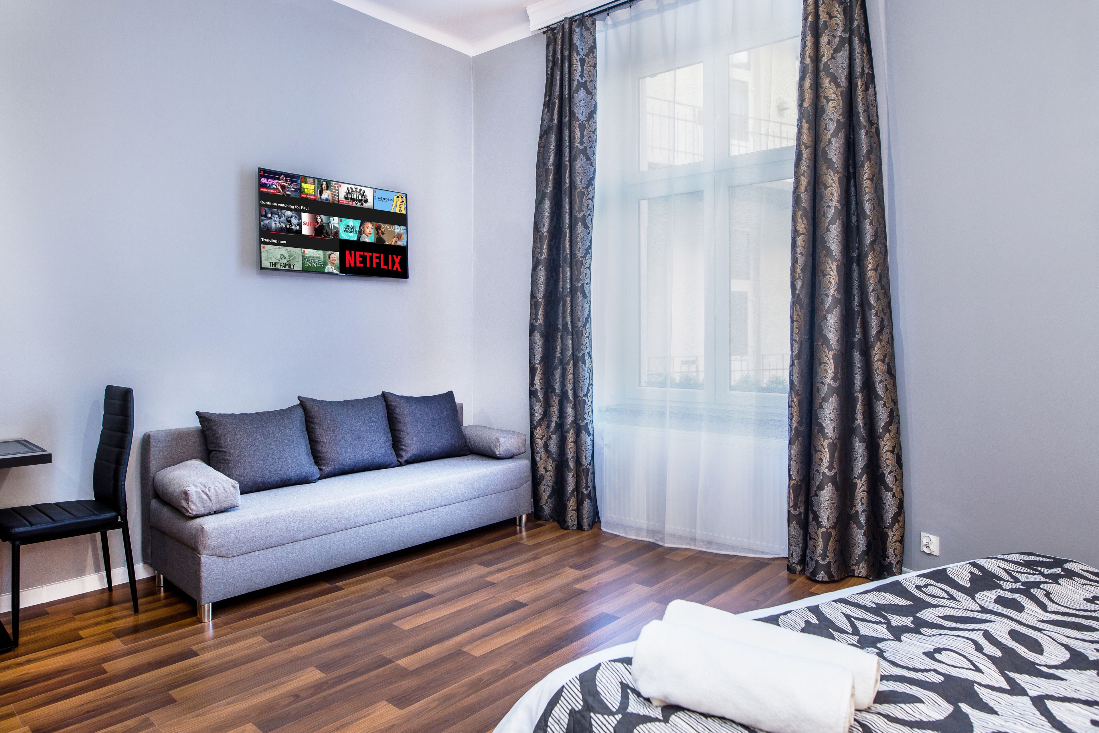 A modern hotel room with a grey sofa, a black chair, a bed with a patterned bedspread, a wall-mounted TV displaying Netflix content, and large windows with sheer curtains.