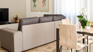 A modern living room with a sofa, a TV, a dining table with chairs, and a hanging light fixture.