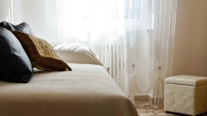 A hotel room with a bed, a small white ottoman, sheer curtains, and a view of the outside.