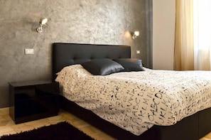 A modern bedroom with a dark headboard, a patterned bedspread, and two wall-mounted lights.