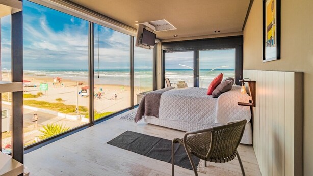 A bedroom with a large window overlooking a beach, a bed with pillows, a chair, and a mounted TV. - Baluarte Pousada (Torres)