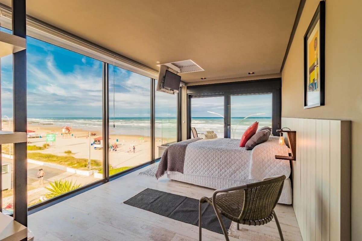 A bedroom with a large window overlooking a beach, a bed with pillows, a chair, and a mounted TV.