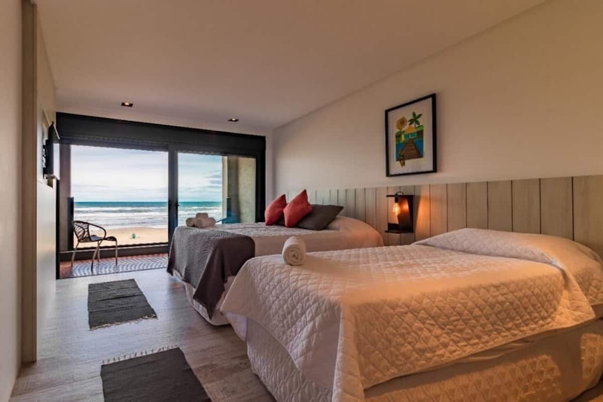 A bedroom with two beds, a large window with a view of the beach, and a framed picture on the wall.