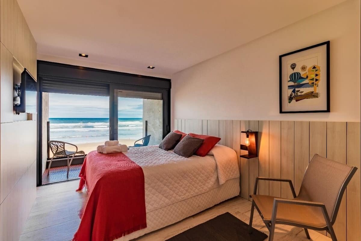 A modern bedroom with a large bed, a view of the ocean, a framed artwork, and a chair.