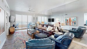 A spacious living room with a large sofa, leather armchairs, a coffee table, and a colorful rug. A large mirror reflects the room, and a television is mounted on the wall. - Beautiful 7th floor condo ON THE BEACH (Orange Beach)