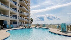 A swimming pool with a curved edge, surrounded by a deck area with lounge chairs and a view of a high-rise building in the background. - Beautiful 7th floor condo ON THE BEACH (Orange Beach)