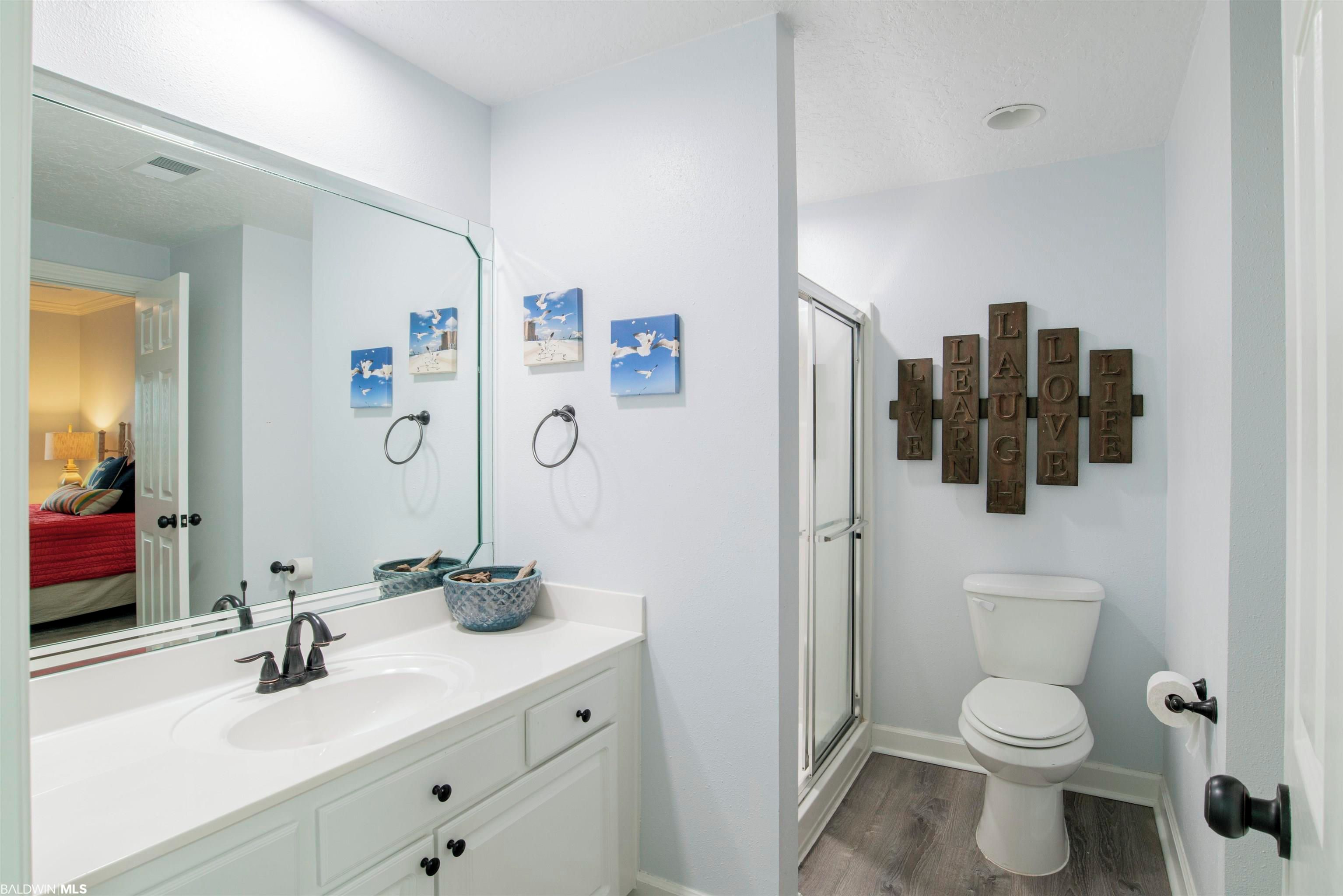 A bathroom with a white sink, a mirror, a toilet, and a shower. There are framed pictures on the wall and a set of wooden plaques with words.