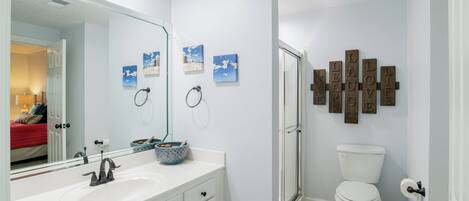 A bathroom with a white sink, a mirror, a toilet, and a shower. There are framed pictures on the wall and a set of wooden plaques with words.