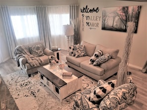 Una sala de estar moderna con un conjunto de sofás, una mesa de centro y almohadones decorativos. Un cartel en la pared dice "welcome to Valley Manor".