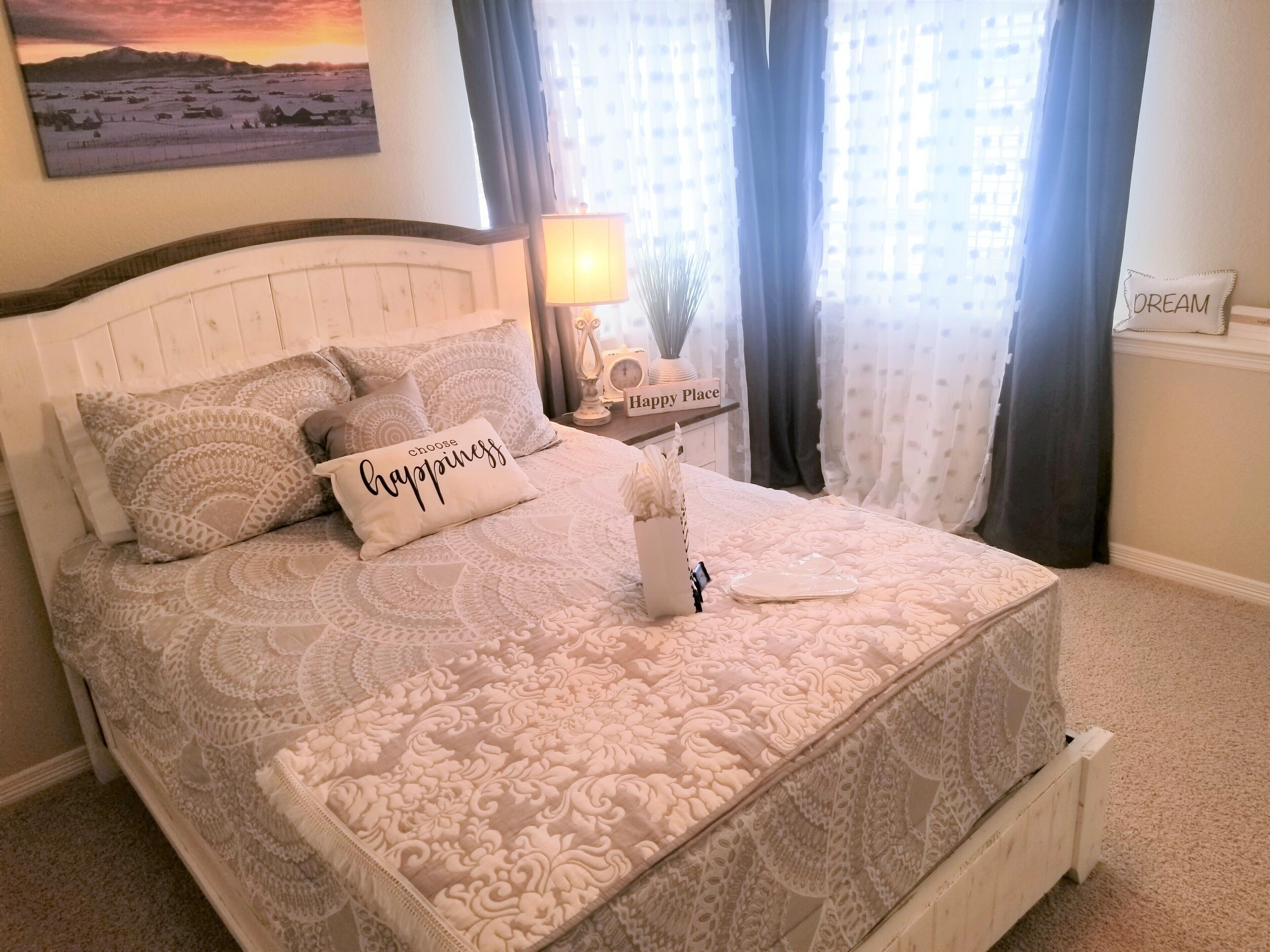 Een net opgemaakt bed met een bedsprei en kussens, een nachtkastje met een lamp, een aan de muur hangend schilderij, en een bordje met de tekst 'DREAM'.