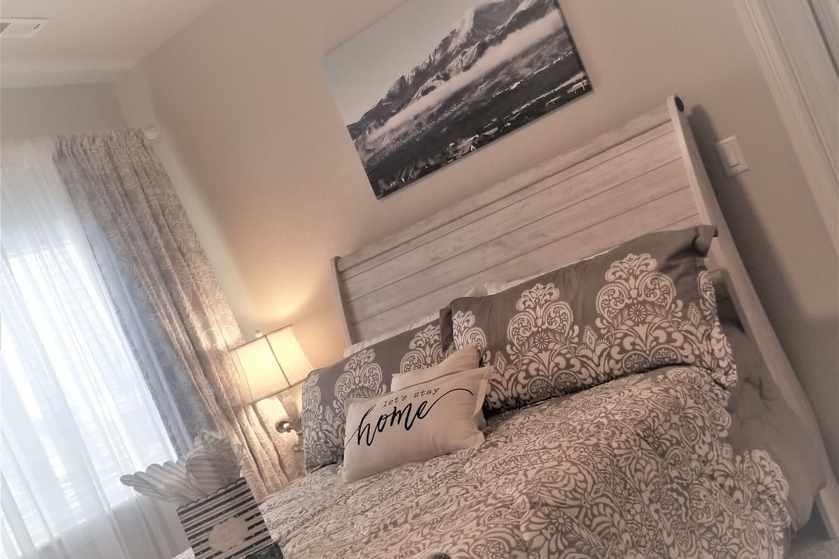A neatly arranged bedroom with a bed, a bedside lamp, a framed picture on the wall, and a window with curtains.