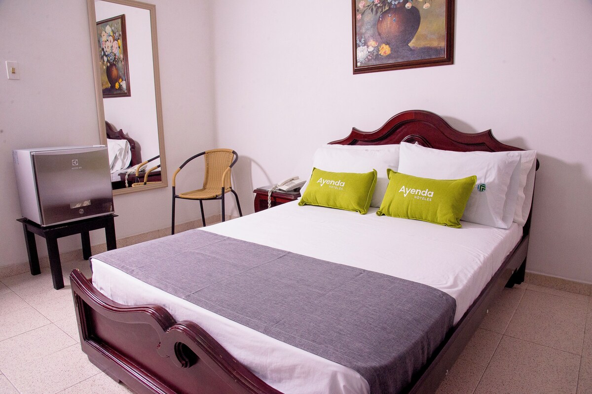 A neatly made bed with a grey bedspread and two pillows, one green and one white, in a hotel room with a wooden headboard.