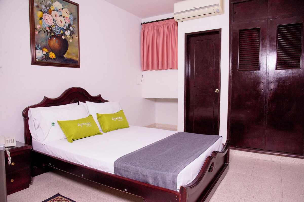 Una cama bien hecha con sábanas blancas y dos almohadas, una mesita de noche de madera con un teléfono, un cuadro de flores en la pared, una cortina rosa y una puerta de madera oscura.
