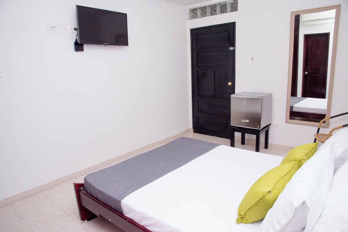 A bedroom with a bed, a television mounted on the wall, a door, and a small table with a heater.