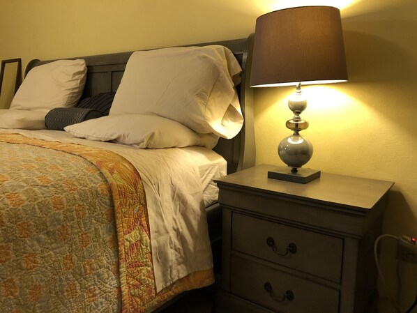 Una cama bien hecha con colcha, almohadas y una lámpara de noche sobre una mesita de noche con cajones.