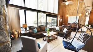 A spacious living room with a large sofa, a coffee table, and a hanging chair. There are large windows providing natural light and a view of the outdoors. - On Lake, WOW Views! 4 Kings, Big Deck. Walk to Marina, Concerts, Events (Dillon)
