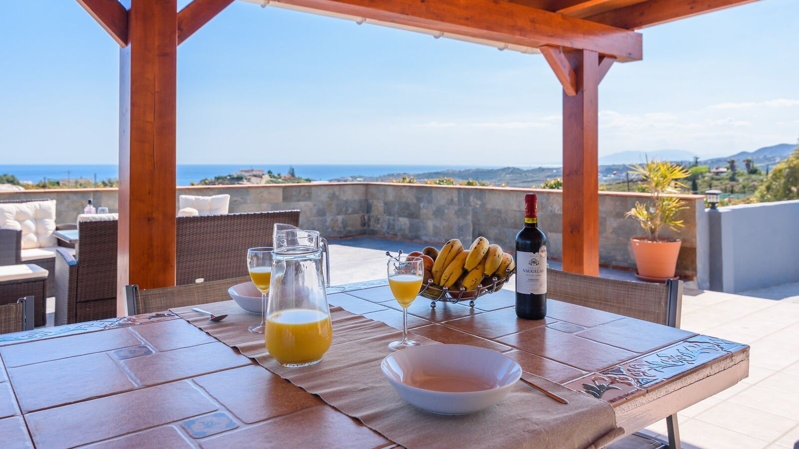 A table set for breakfast with a view of the sea, a bottle of wine, and a bowl of fruit.