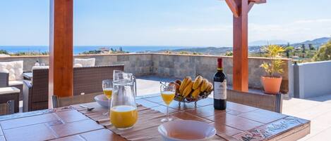 A table set for breakfast with a view of the sea, a bottle of wine, and a bowl of fruit.
