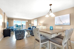 A spacious dining area with a wooden table and white chairs, a balcony with blue chairs, and a painting of sailboats on the wall. - Sandy Key 721: 3 BR/2 BA On-Site Rental Office, Perdido Key Beachfront Condo, Sleeps 8 (Pensacola)