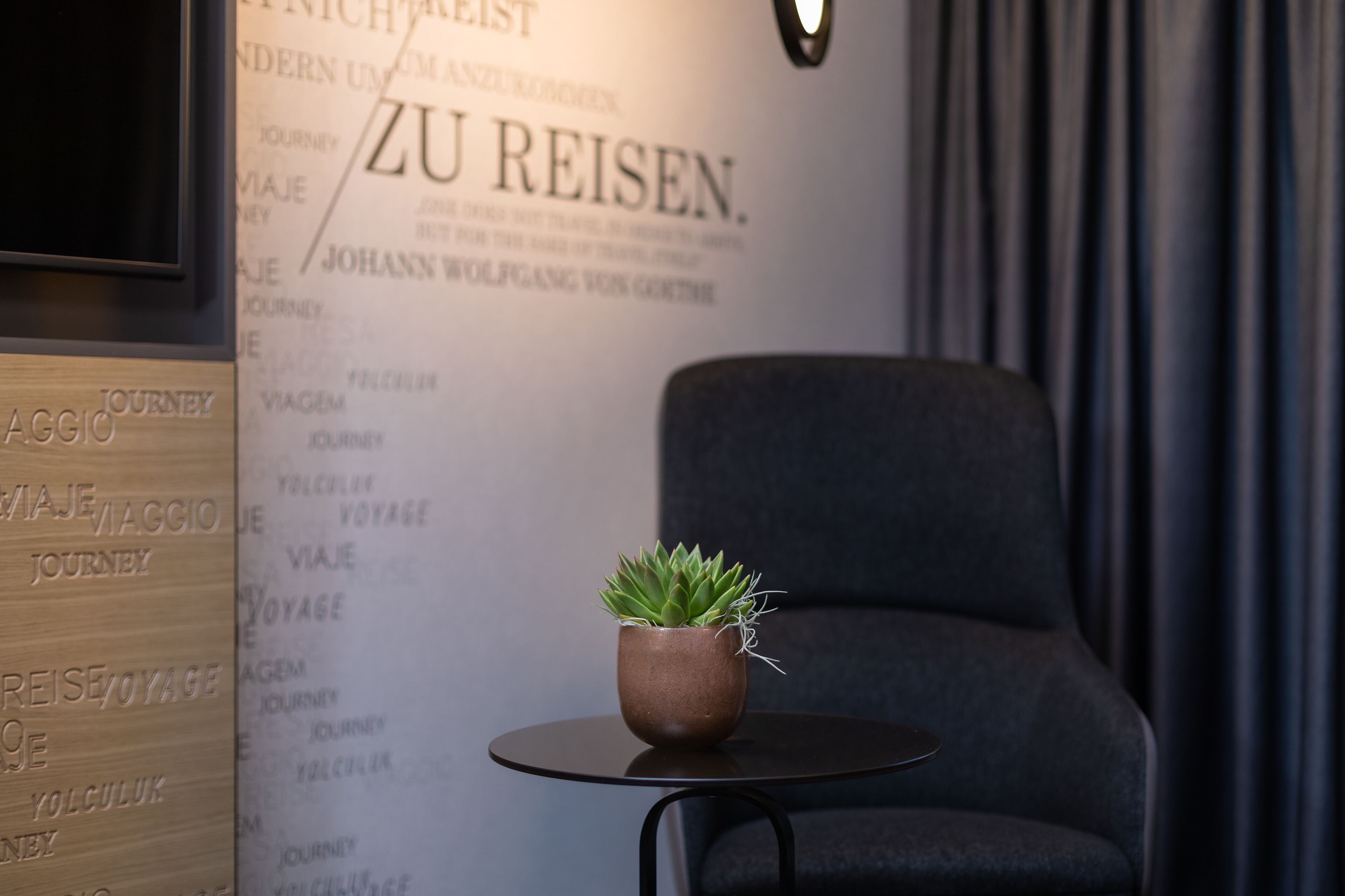 Une pièce moderne avec un fauteuil gris, une petite table sur laquelle est posée une plante en pot, et un mur orné d’un texte rédigé en plusieurs langues.