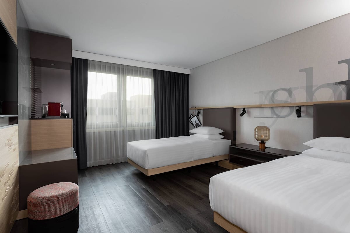 A modern hotel room with two single beds, a small seating area, a desk, and a large window with curtains.