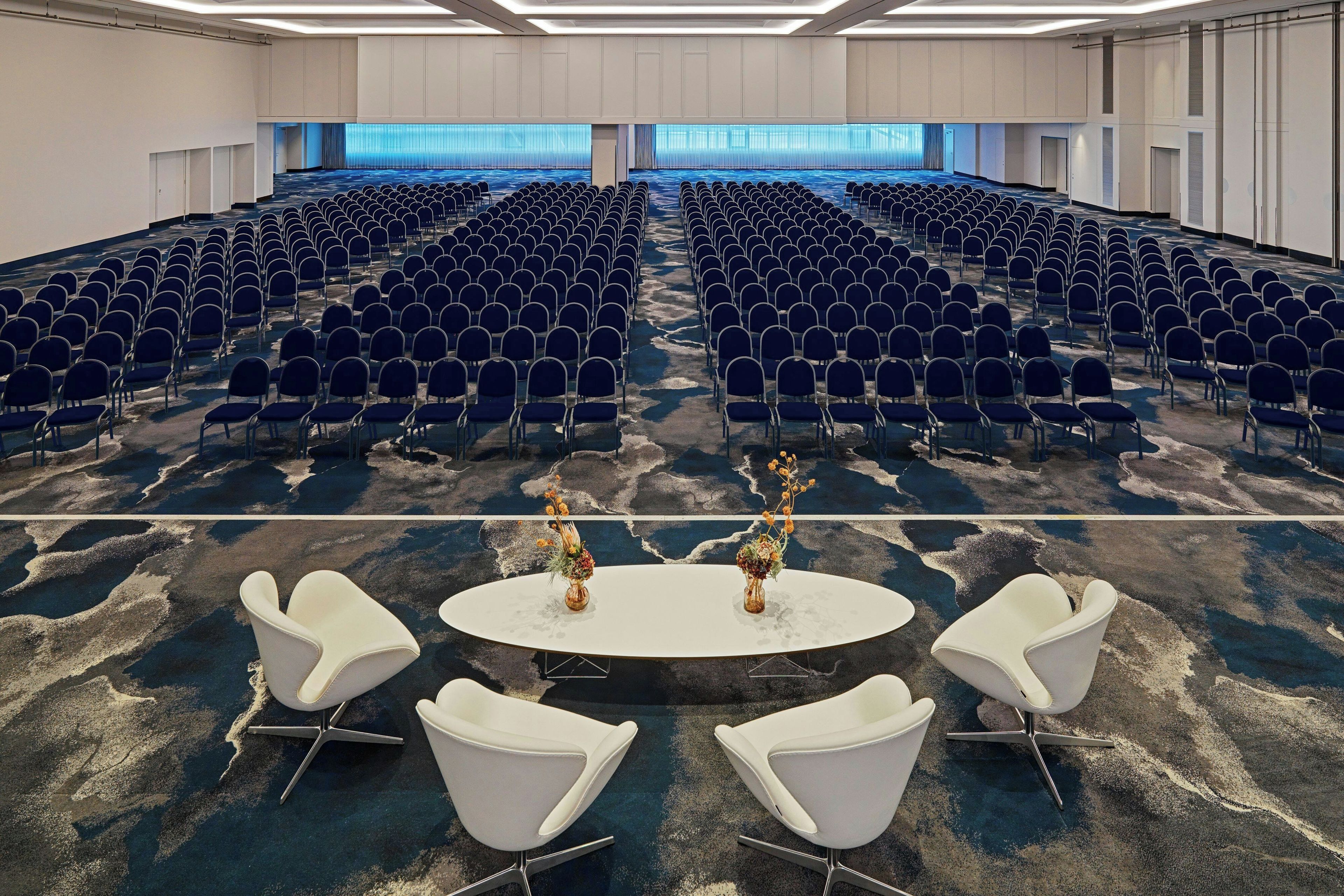 A conference room with rows of blue chairs, a white table with a vase of flowers, and a carpeted floor.