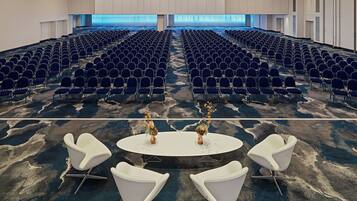 A conference room with rows of blue chairs, a white table with a vase of flowers, and a carpeted floor.