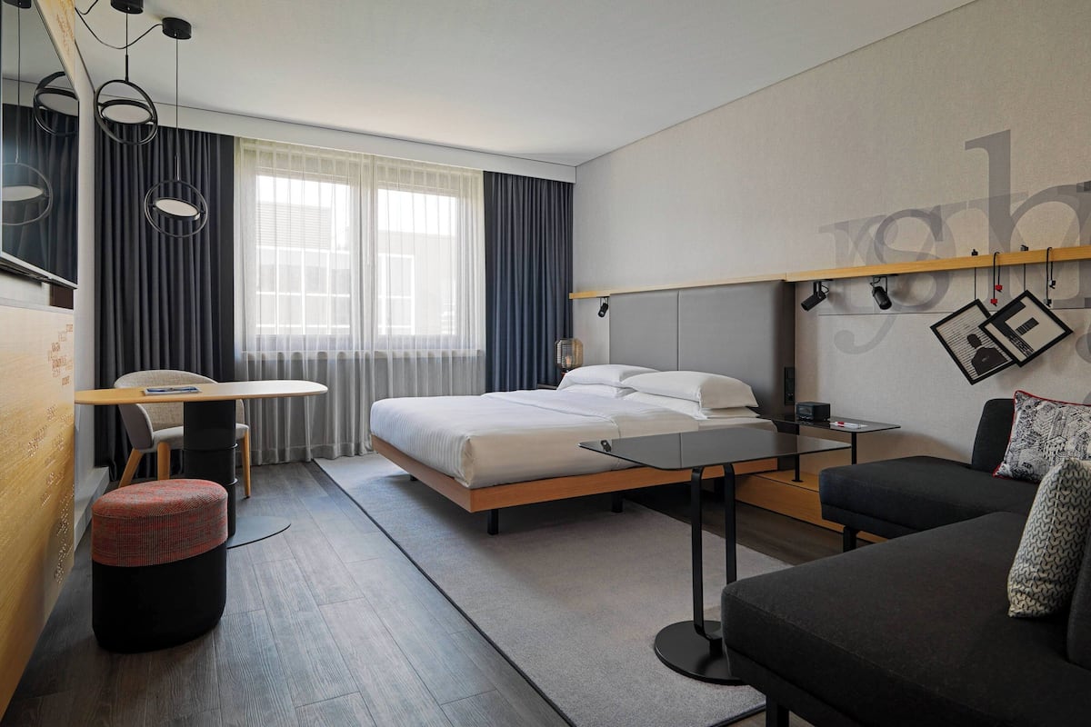 A modern hotel room with a bed, a desk, a sofa, and a large window with curtains.