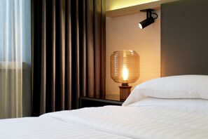 Un lit bien fait, avec du linge de lit blanc, une lampe moderne dotée d’un abat-jour cylindrique et des rideaux foncés à une fenêtre.