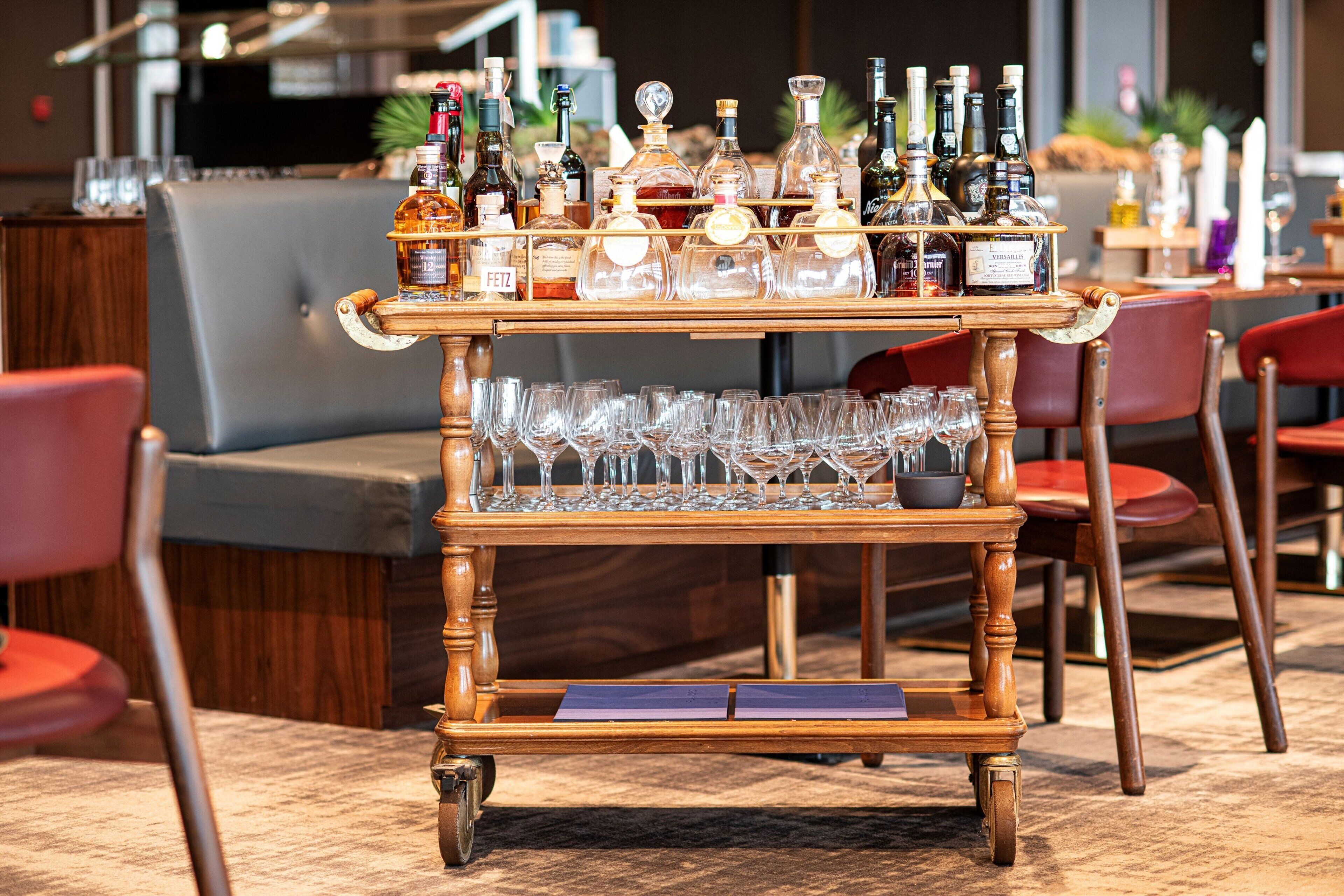 A bar cart with various bottles and glasses, placed in a room with red chairs and a grey sofa.