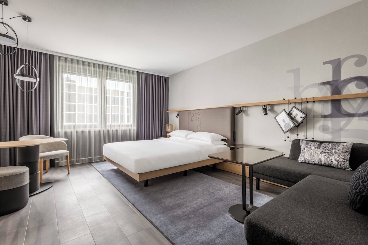 A modern hotel room with a bed, a desk, a sofa, and a large window with curtains.