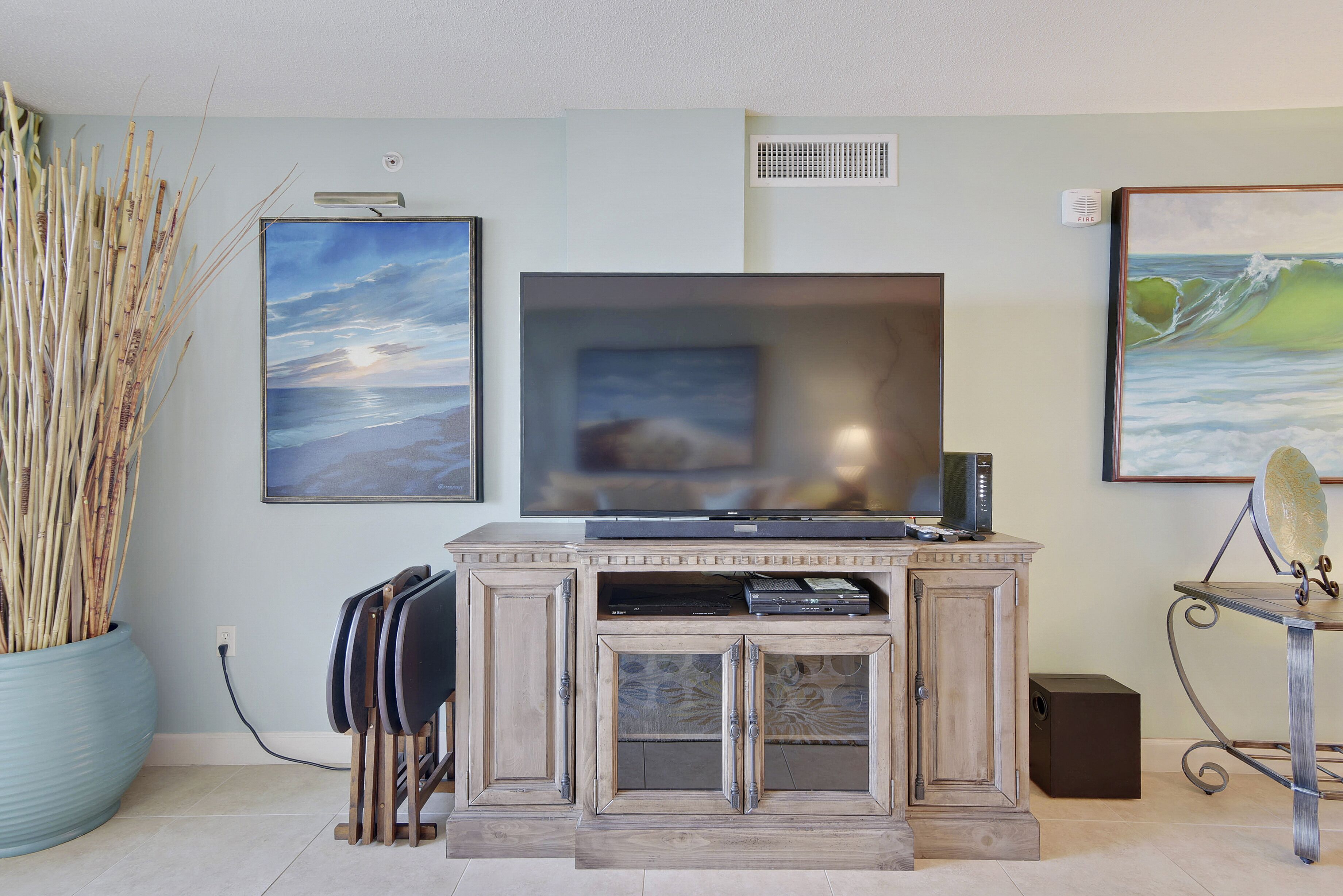 A living room with a flat-screen TV mounted on a wooden cabinet, a large potted plant, and two framed artworks on the wall.