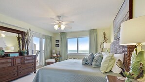 Un dormitorio con una cama grande, un aparador, un ventilador de techo y vistas al océano a través de las ventanas.