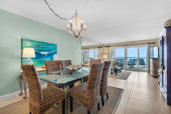 A dining area with wicker chairs and a glass table, a chandelier, and a painting of a seascape.