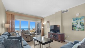 Ein geräumiges Wohnzimmer mit einer großen Couch, einem Couchtisch und einem Flachbildfernseher. Eine Glastür öffnet sich zu einem Balkon mit Blick auf den Ozean.