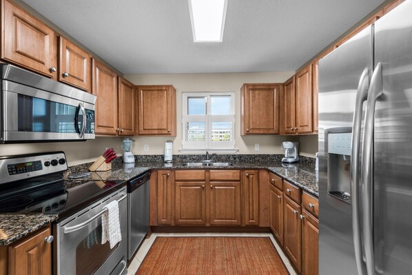 A modern kitchen with wooden cabinets, stainless steel appliances, a granite countertop, and a window with blinds. - Sandy Key 312: 2 BR/2 BA Beachfront Condo in Perdido Key, FL On-Site Rental Office, Sleeps 6 (Pensacola)