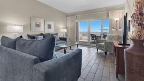 A modern living room with grey sofas, a glass coffee table, and a TV on a wooden stand. There is a balcony with a pool view. - Sandy Key 126: 2 BR/2 BA Beachfront Condo, Perdido Key, FL. On-Site Rental Office, Sleeps 6 (Pensacola)