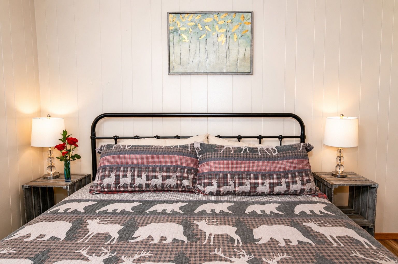 Ein Bett mit einer karierten Decke mit Tiermotiven, das von zwei Nachttischen mit Lampen und einer Blumenvase flankiert wird.