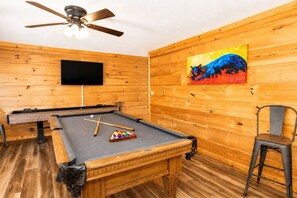 Een biljart met een keu en ballen, een aan de muur bevestigde televisie, en een schilderij aan de wand.