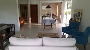 Un salon avec un canapé blanc, un fauteuil bleu et une table basse en bois. On aperçoit à l’arrière-plan un coin repas avec une table et des chaises.