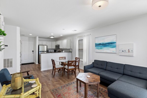 A modern living room with a grey sofa, wooden dining table, and chairs, a kitchen in the background, and artwork on the walls.