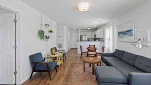 A modern living room with a grey sofa, wooden coffee table, and blue armchairs. A kitchen is visible in the background with white cabinets and a stainless steel refrigerator. - Perfect Year-Round! 2nd floor golf course condo, pool & hot tub, close to beach (Pawleys Island)