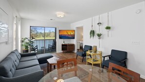 A modern living room with a sofa, chairs, a glass coffee table, a TV, and a balcony with a view. - Perfect Year-Round! 2nd floor golf course condo, pool & hot tub, close to beach (Pawleys Island)