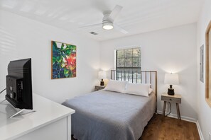 A bedroom with a bed, a desk with a TV, a ceiling fan, a window, and two bedside lamps.